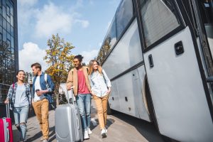 young smiling interracial couples with wheeled bags walking near travel bus at city street